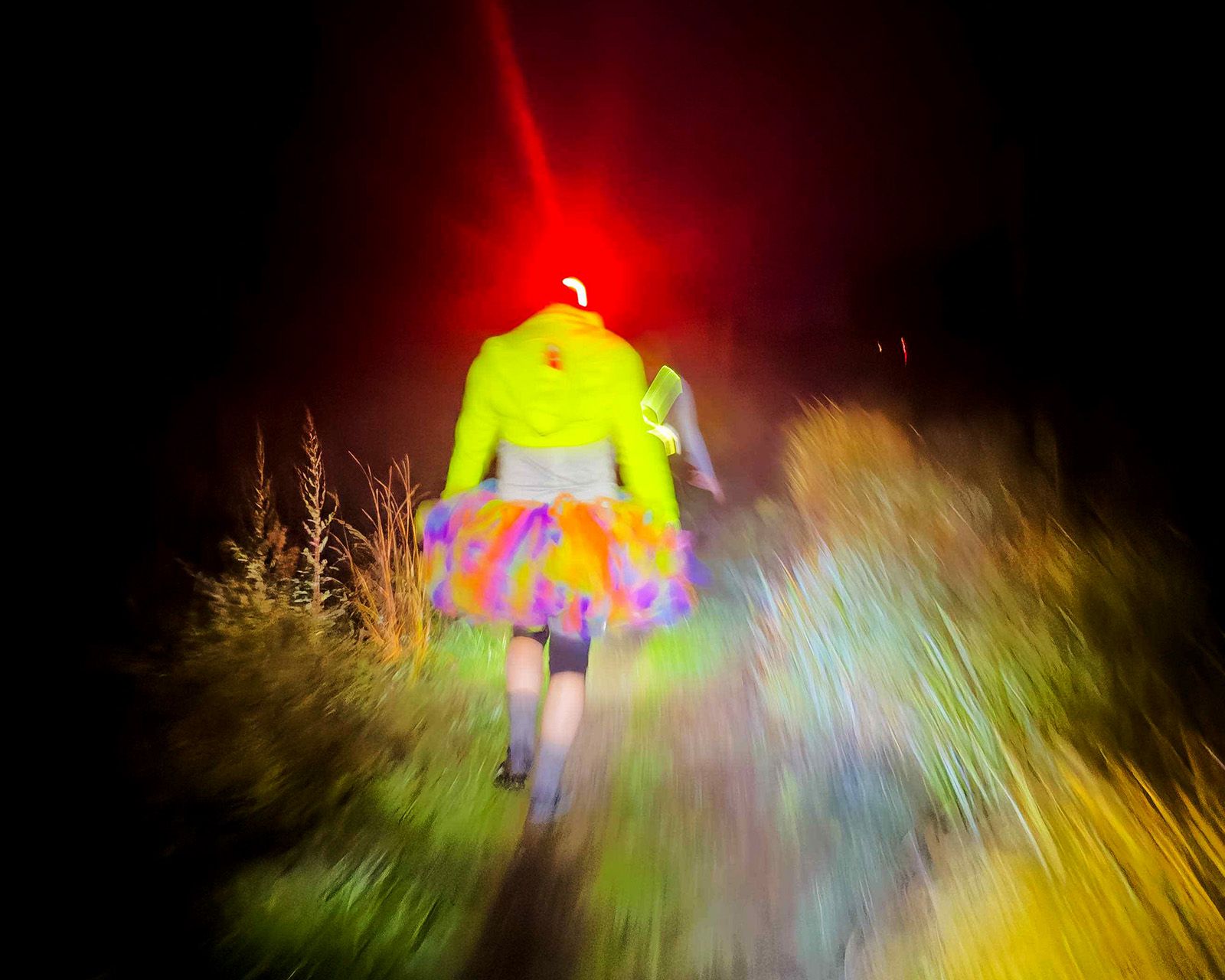 A hasher in a neon jacket and rainbow tutu on a night trail